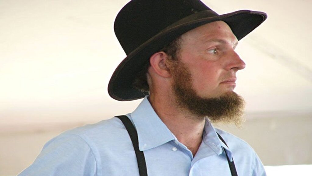 An Amish man with beard wearing a hat and blue blouse.